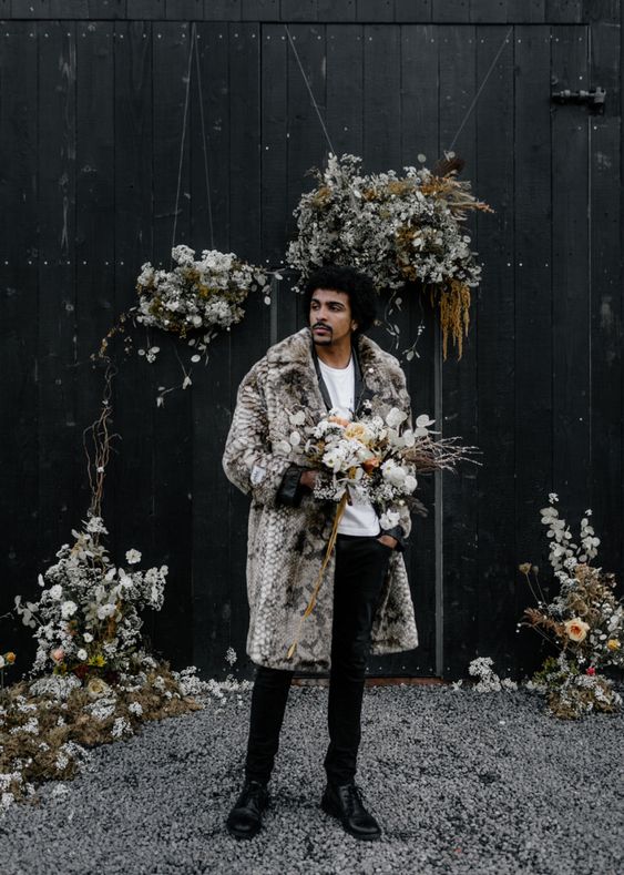 a modern groom's look with a black velvet pantsuit, a white t-shirt, black shoes and a greige faux fur coat with a print is amazing for a winter affair