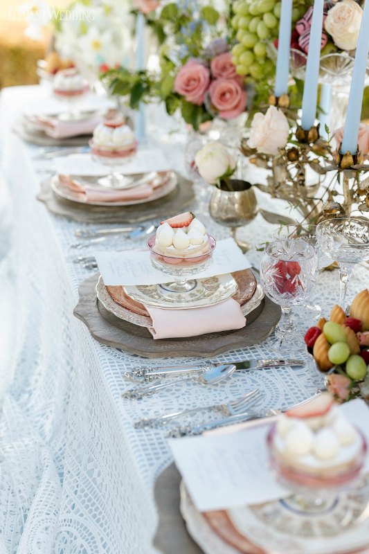 an exquisite wedding tablescape with a lace tablecloth, pastel blue candles, pastel florals and elegant porcelain and delicious sweets