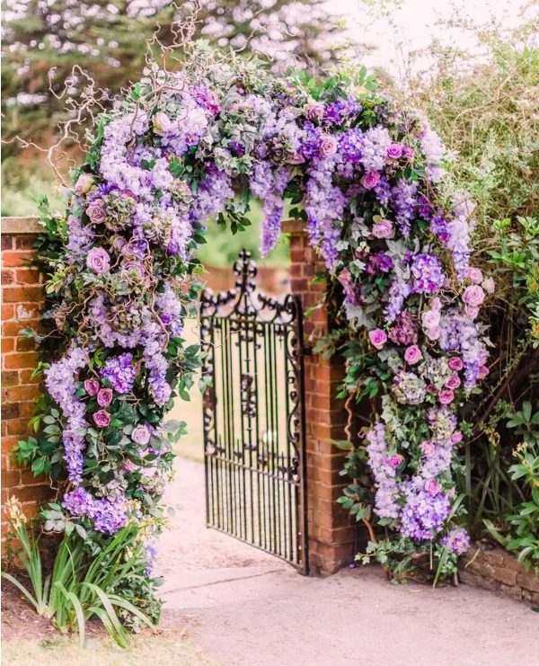 a wedding gate decorated with a gorgeous purple and lilac blooms, vines and greenery is a gorgeous idea for a Bridgerton wedding