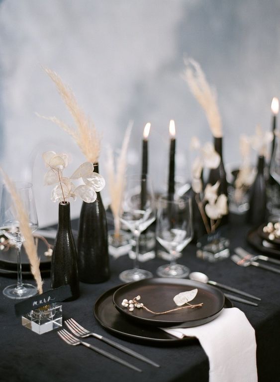a beautiful black and white wedding tablescape with a black tablecloth and white napkins, black vases and candles, dried blooms and grass