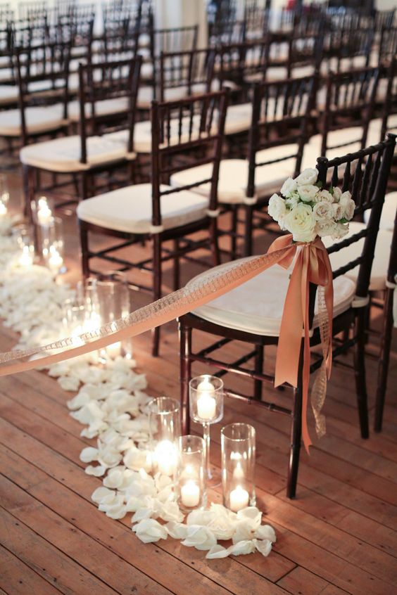 a dark and white wedding chair in the aisle accented with an orange ribbon and a bow plus a white floral arrangement and candles and petals on the floor