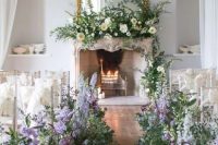 27 a sophisticated wedding ceremony space with a fireplace with candles and lush florals, a crystal chandelier, neutral chairs and greenery and very peri blooms lining up the aisle