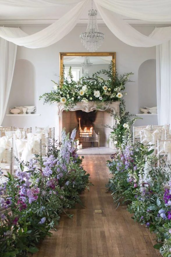a sophisticated wedding ceremony space with a fireplace with candles and lush florals, a crystal chandelier, neutral chairs and greenery and very peri blooms lining up the aisle