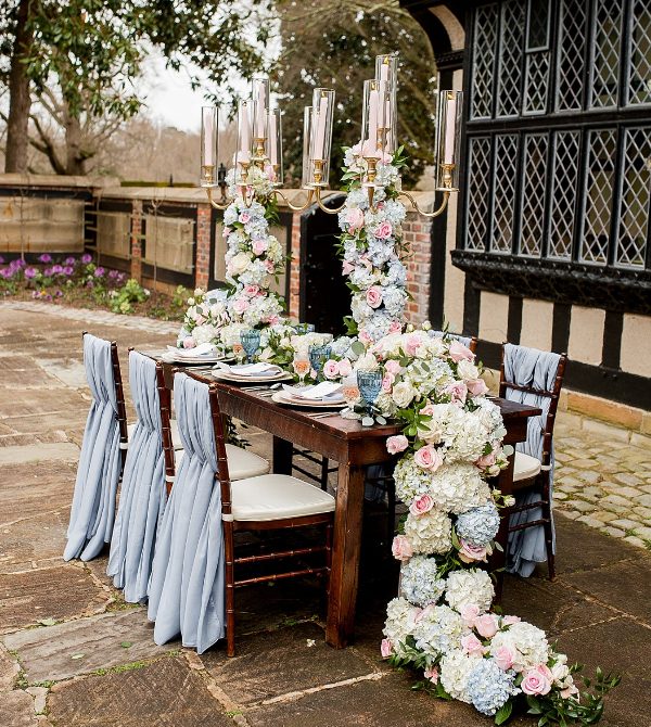 a refined Bridgerton wedding tablescape with tall candelabras decorated with super lush florals, blue and blush glasses, elegant porcelain and pastel blue chair covers