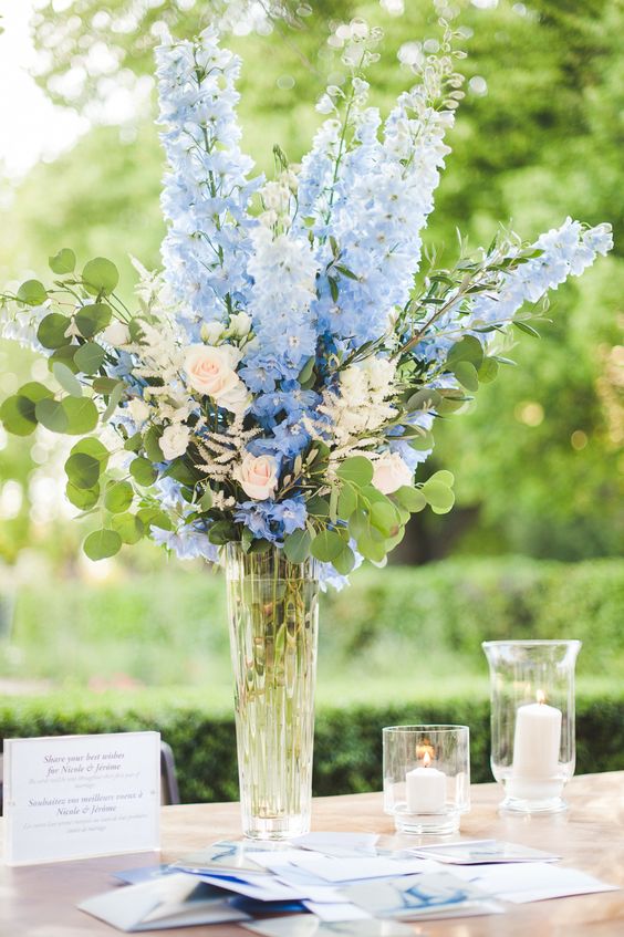 a tall wedding centerpiece of a clear vase with blue and periwinkle blooms plus greenery and candles is a color statement