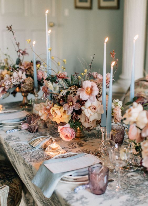a fantastic wedding tablescape with a printed tablecloth, lush blush, peachy and mauve blooms, thin and tall blue candles and napkins