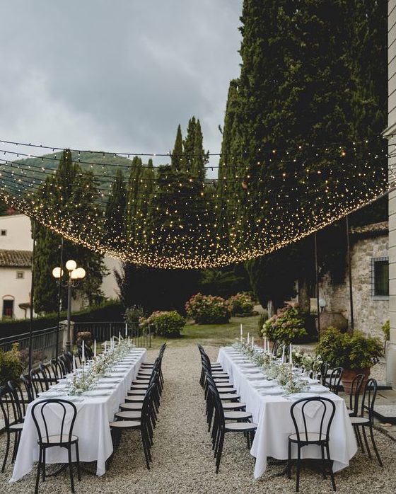 a beautiful and chic al fresco wedding reception with greenery runners and candles, with refined chairs and a warm light canopy