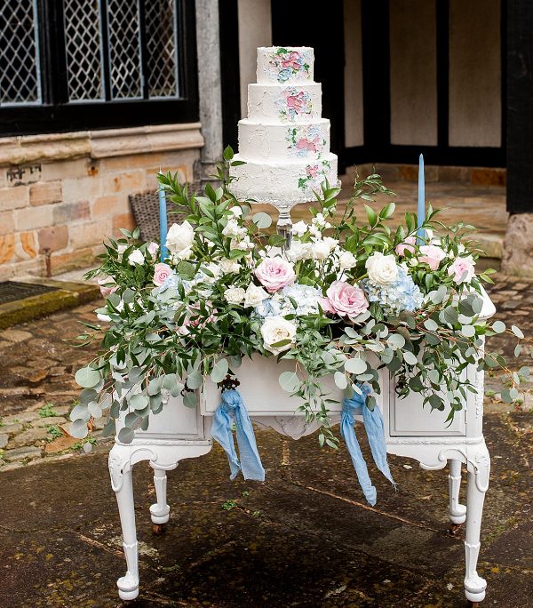 a refined console table decorated with super lush and pretty florals, with blue thin and tall candles, a white textural wedding cake with pasinted blooms