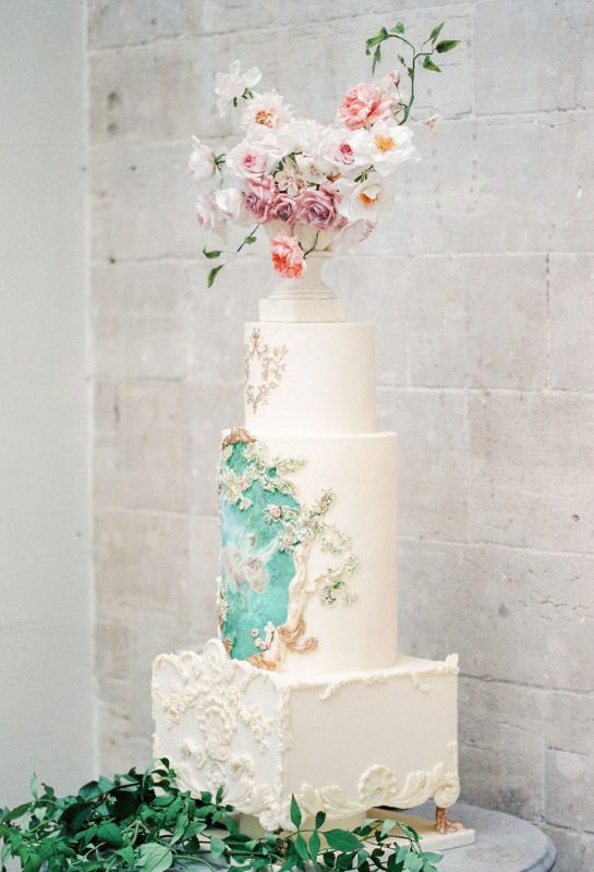 a unique white wedding cake with square and round tiers, with patterns and painting plus an urn with blooms on top