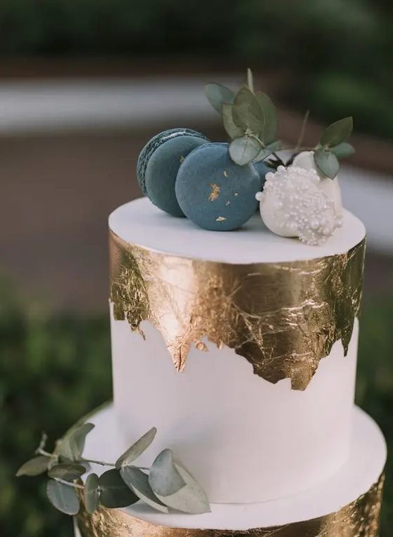 a white wedding cake with gold leaf, blue and white macarons and eucalyptus looks very edgy