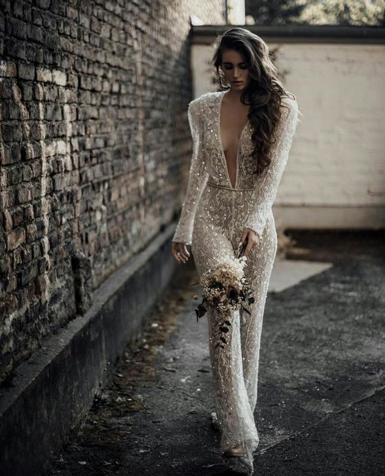 a fully embellished lace bridal jumpsuit with a plunging neckline, long sleeves, accented shoulders and flare pants
