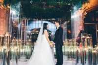 a beautiful lucite wedding arbor topped with lush greenery and tall glasses with floating candles for a garden feel at an indoor ceremony