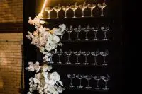 a black metal champagne wall with elegant glasses, white floral arrangements and a neon sign on top is amazing