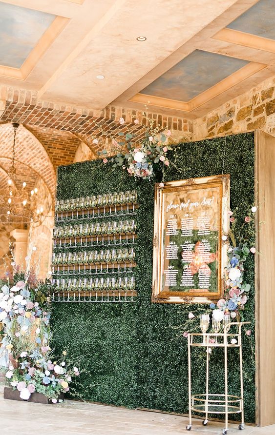 a boxwood wall with a seating chart on a mirror, wooden holders for glasses and some pastel floral arrangements