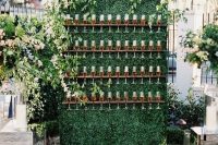 a boxwood wall with wooden flute holders, lush greenery and neutral blooms on top and pillar candles in glasses