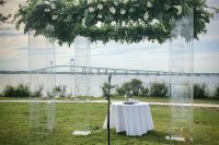 a clear lucite wedding arbor covered with lush greenery and white blooms on top plus a sea and a bridge view in the backdrop