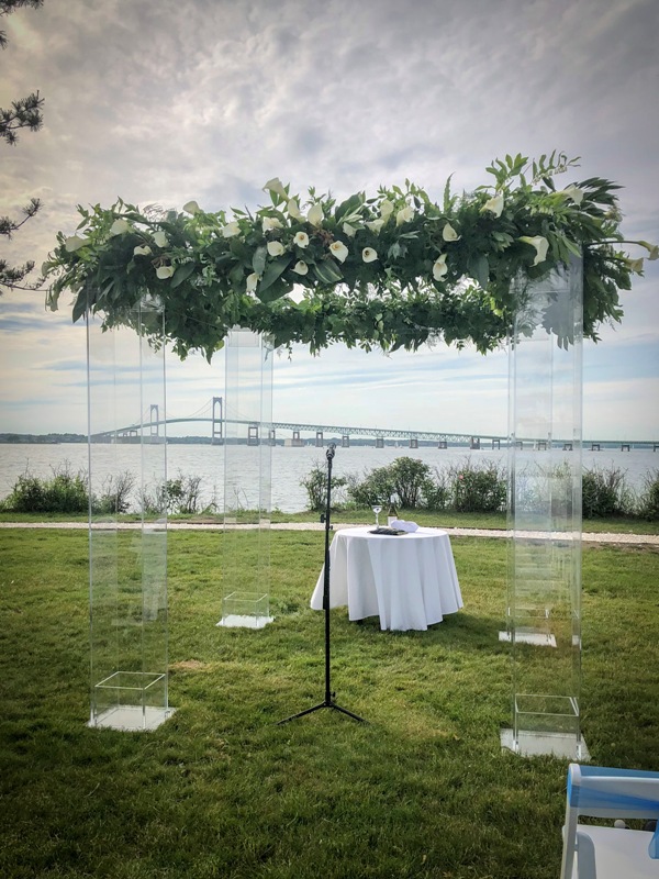 a clear lucite wedding arbor covered with lush greenery and white blooms on top plus a sea and a bridge view in the backdrop