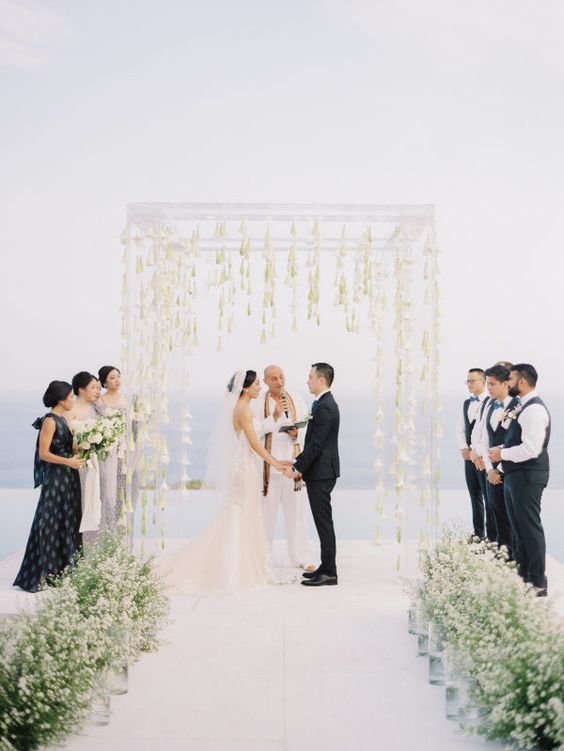 a clear lucite wedding arch with white blooms hanging down from it that seem floating in the mid-air plus a sea view