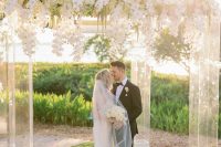 a large acrylic wedding arbor covered with lush white orchids and with a view of a field as a backdrop