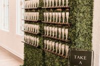 a large boxwood champagne wall with wooden glass stands and a black sign next to it is a cool idea