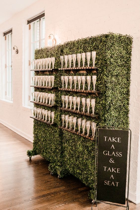 a large boxwood champagne wall with wooden glass stands and a black sign next to it is a cool idea