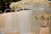 a large white platform with an acrylic wedding arbor topped with white orchids and with orchid arrangements suspended around plus pillar candles