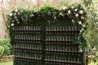 a living greenery wall with wooden champagne glass holders and lots of greenery and neutral blooms covering the top of the wall