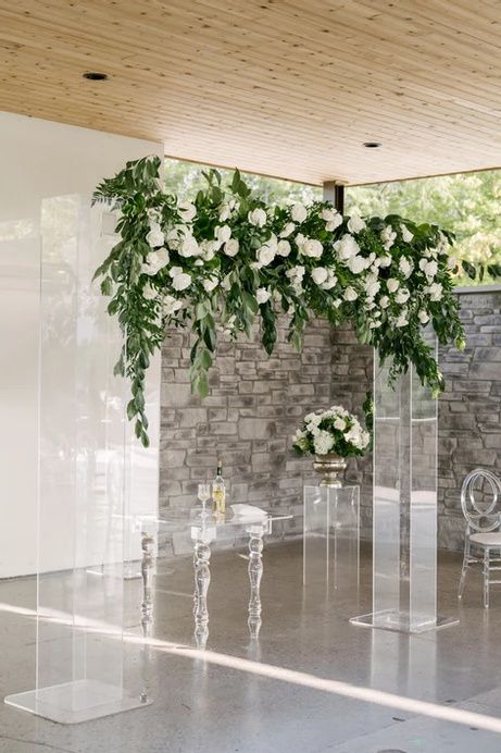 a lucite wedding arch decorated with greenery and white blooms on top is a very chic and beautiful idea with a strong modern feel