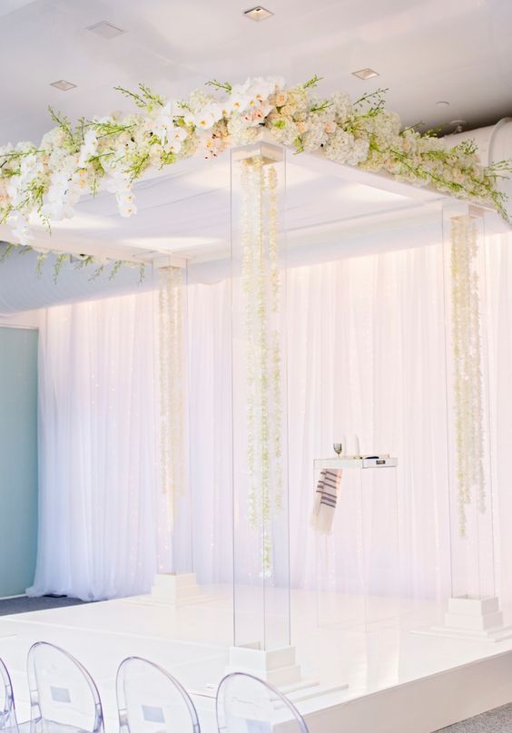 a lucite wedding chuppah with floral petals inside the pillars and lush white florals on top is a lovely idea for a modern luxurious wedding