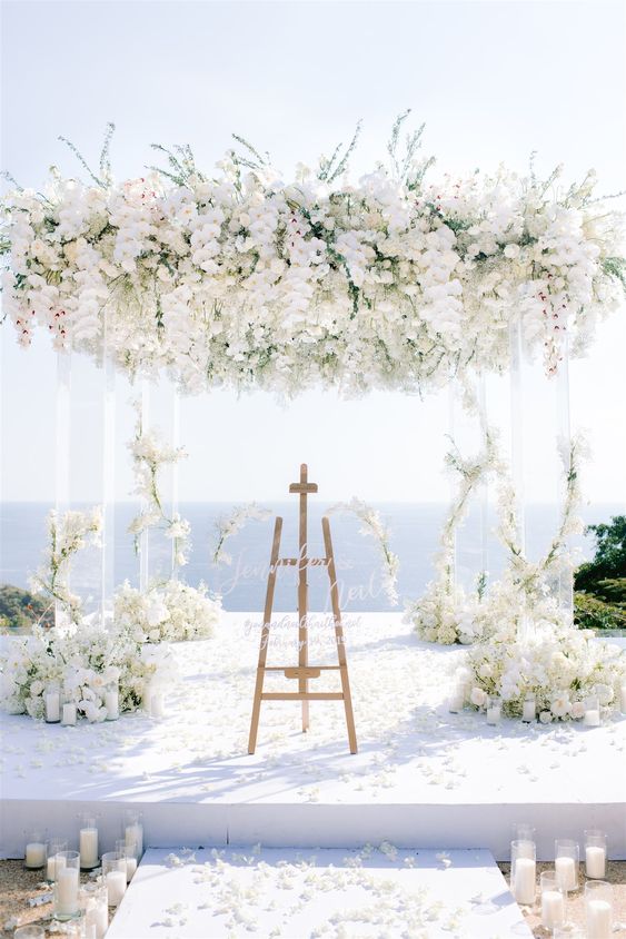 a luxurious wedding ceremony space with a sea view, an acrylic arbor with white blooms covering its top and the pillars, pillar candles