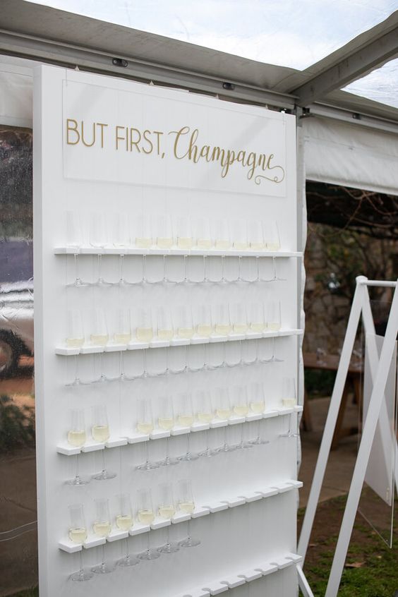 a minimalist white champagne wall with holders for glasses and an acrylic sign with a bit of gold calligraphy is cool