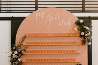 a peachy curved champagne wall with several rows of glasses, neutral and bold floral arrangements with greenery and stars and calligraphy