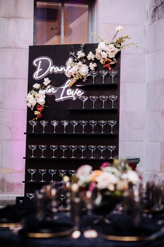 a refined black champagne wall with a neon sign and lovely pink and orange blooms and greenery is a gorgeous solution