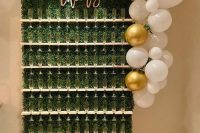 a small boxwood champagne wall with neutral holders, calligraphy, white and gold balloons with greenery on top