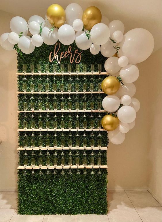 a small boxwood champagne wall with neutral holders, calligraphy, white and gold balloons with greenery on top