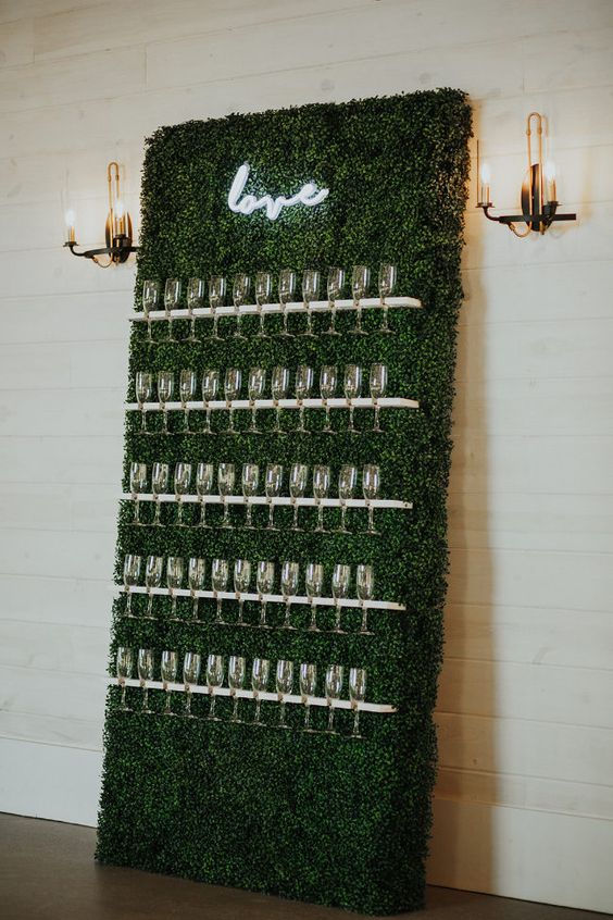 a small boxwood wall with white flute holders and a small neon sign is a cool idea for a modern and not very big wedding