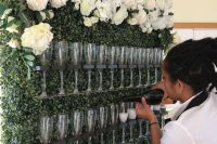 a stylish wedding champagne wall covered with boxwood, with dark stained wooden holders and lush white florals on top the wall