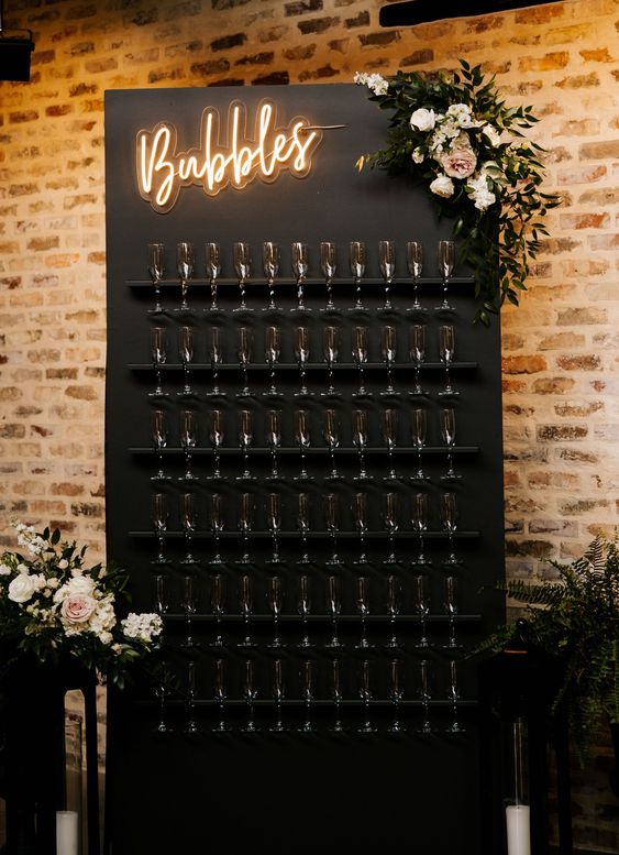 a super stylish black champagne wall with matching holders, a neon sign and a delicate pastel floral arrangement on the corner