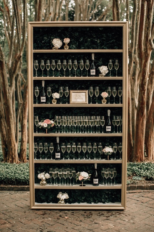 a unique champagne glass wall - a bookcase with greenery, glasses and bottles plus pastel bloom arrangements
