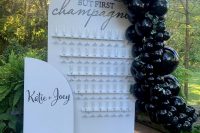 a white curved champagne wall with neutral flute holders, calligraphy, greenery on top, a bold blakc balloon garland covering one side