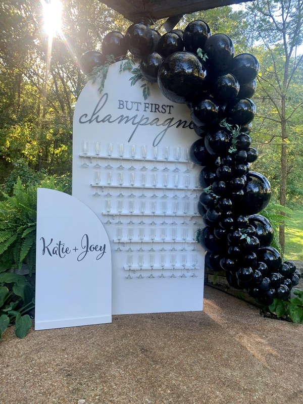 a white curved champagne wall with neutral flute holders, calligraphy, greenery on top, a bold blakc balloon garland covering one side