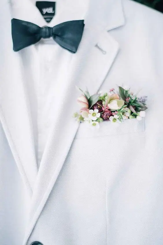 a white tux with a pocket square done with hellebores, wax flower and tight red buds plus greenery for a slight pastel touch