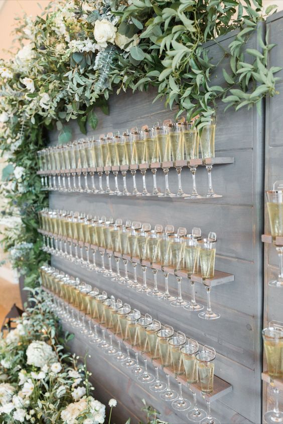 a whitewashed plywood wall with maching flute holders, a lush greenery and white flower garland on top, glasses as escort cards