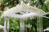 an exquisite modern clear acrylic wedding arbor with lush white blooms on top and a cool jungle view is amazing