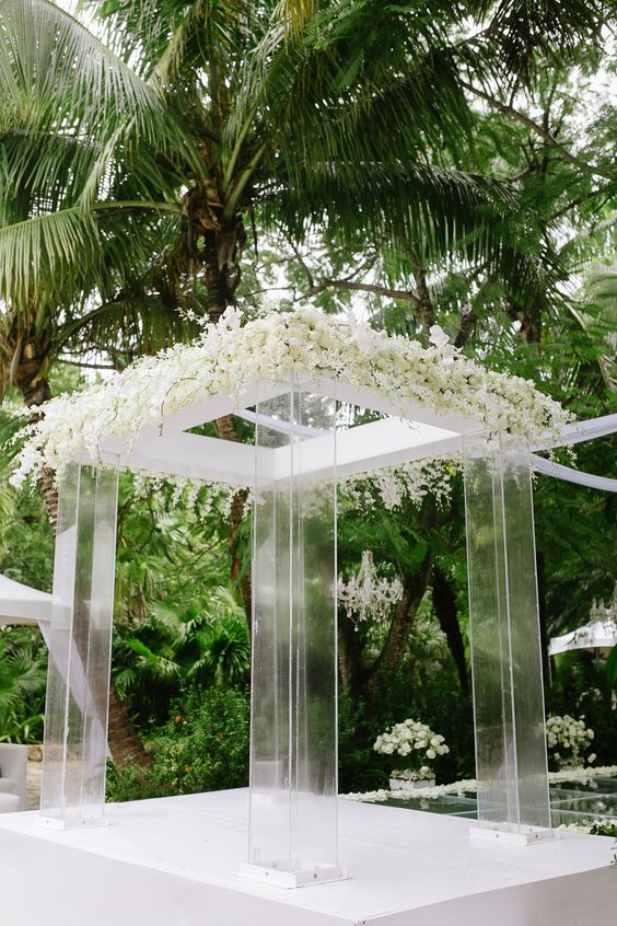 an exquisite modern clear acrylic wedding arbor with lush white blooms on top and a cool jungle view is amazing