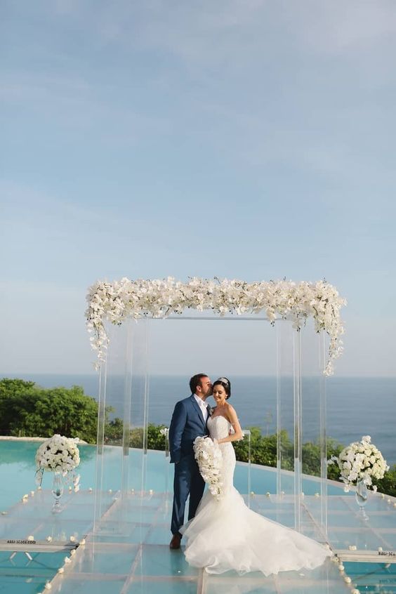 an exquisite wedding ceremony space with a sea view, a clear platform with white floral arrangements, a clear acrylic arch topped with blooms