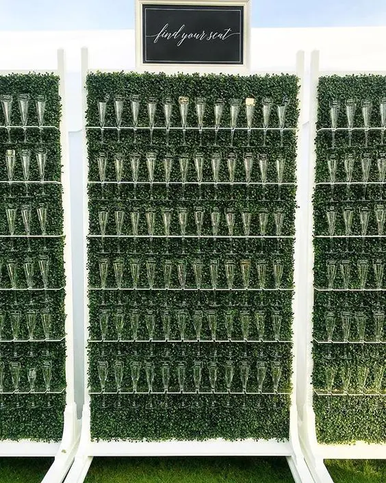 an oversized boxwood wall with white flute holders and a black sign is a cool idea for a big wedding