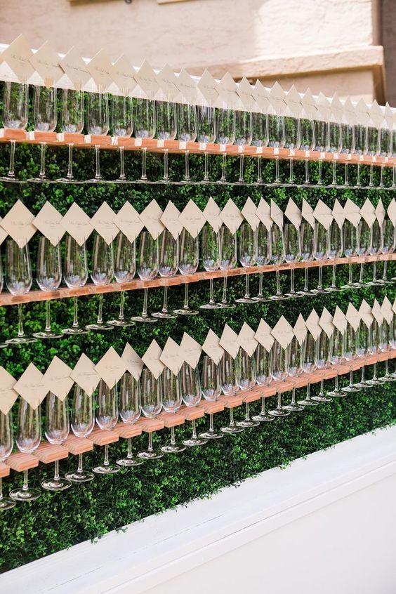 an oversized greenery wall with wooden holders for champgane flutes and escort cards on top each flute is amazing