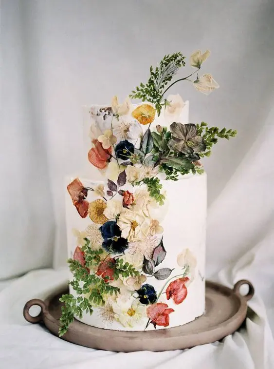 a sophisticated white buttercream wedding cake with presed dried flowers and some fresh blooms and leaves attached to the cake