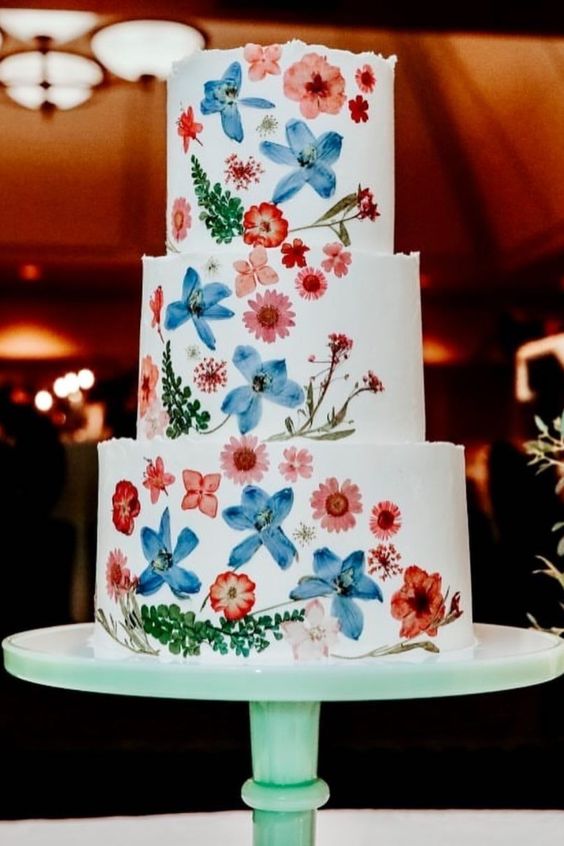 a white buttercream wedding cake with uneven edges and red and blue pressed flowers and leaves to form a pattern and a color scheme